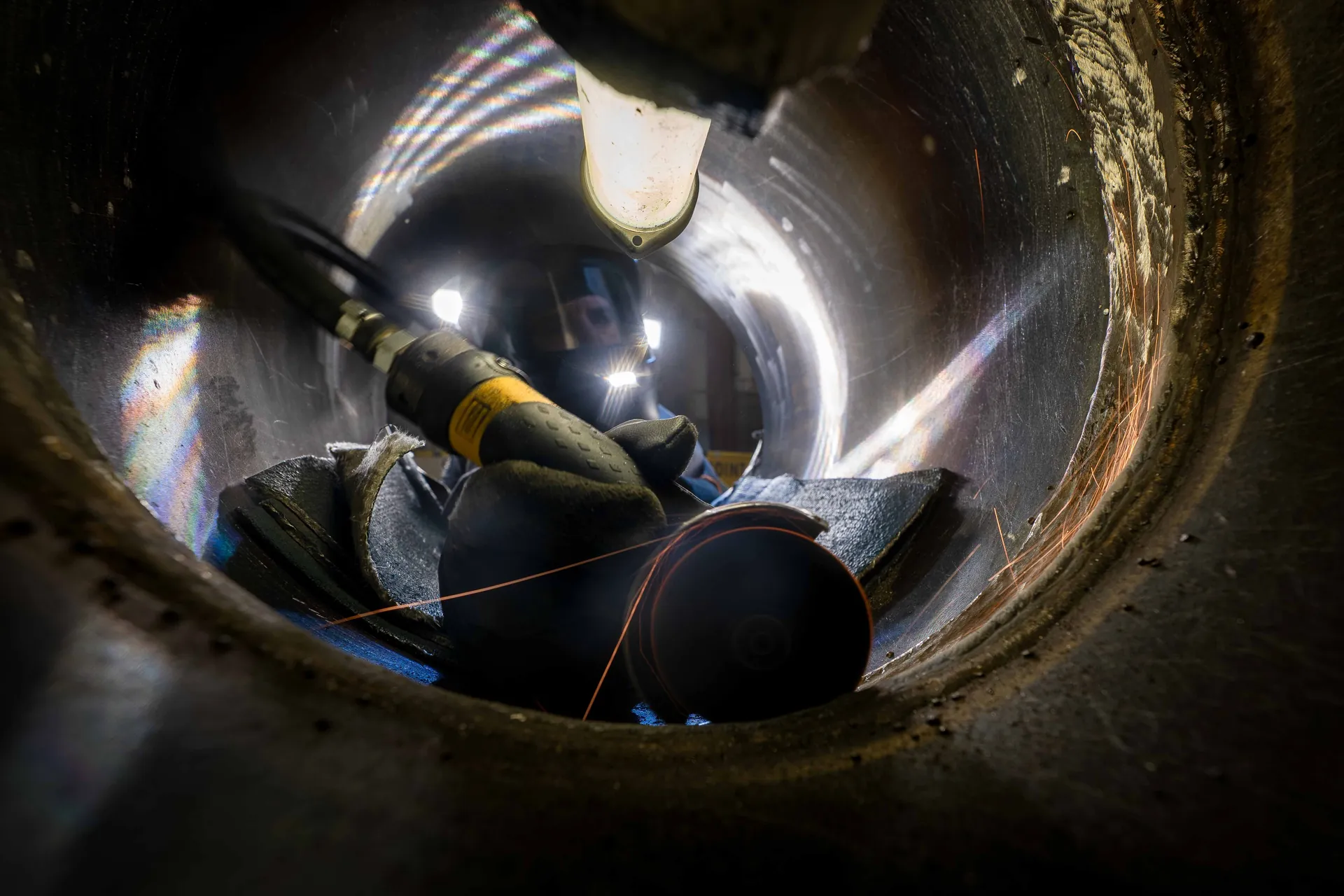 Technicien meulant l'intérieur d'une tuyauterie circulaire, reflets irisés et étincelles