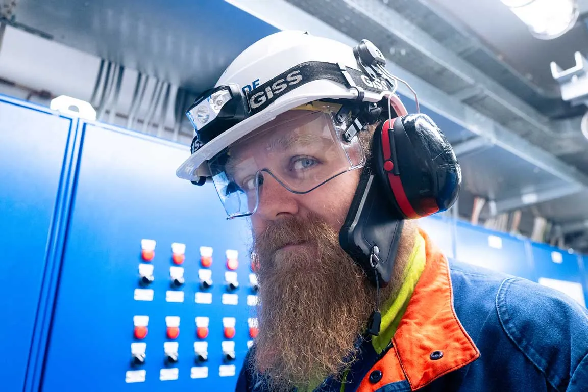 Portrait d'un technicien barbu avec casque GISS, visière et antibruit, devant un tableau électrique bleu