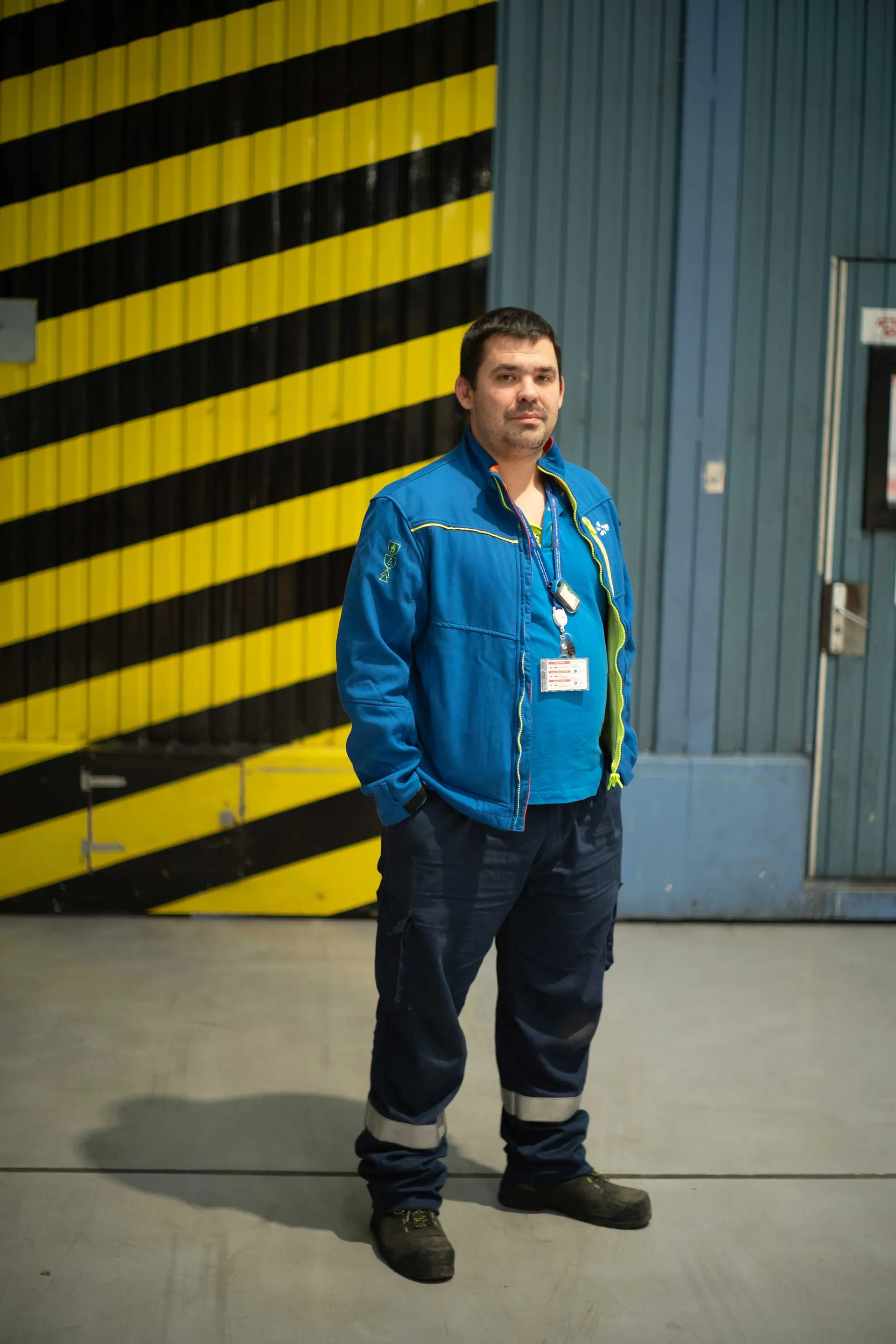 Technicien EDF en combinaison bleue posant devant une porte de hangar à bandes jaunes et noires