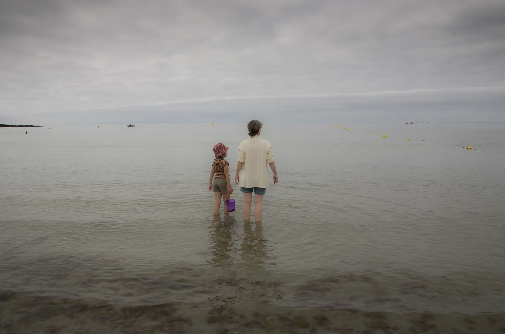 Adulte et enfant debout dans l'eau peu profonde de dos, seau violet, ciel couvert